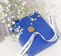 A blue gift with a white ribbon and a gold-colored seal, resting on a surface with a white flower in the background.