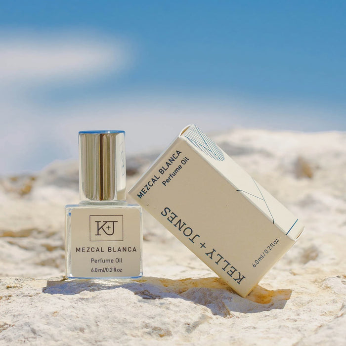 Kelly + Jones Mezcal Blanca perfume oil bottle and packaging on a sandy beach surface with a blue sky.